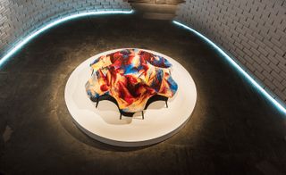 View of a 'The Nature Of Motion’ installation featuring a colourful communal seat by Sebastian Wrong on a circular plinth against a white curved brick wall and dark flooring