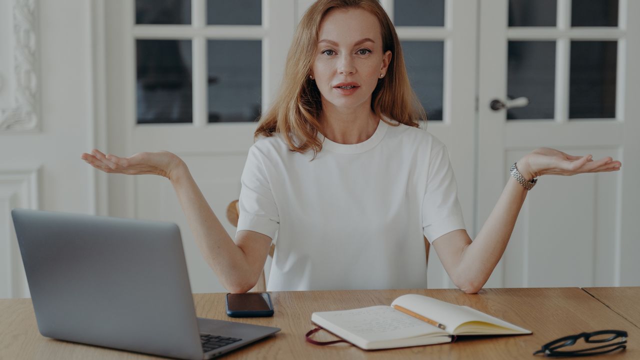 A woman puts her hands palms up as if weighing choices while sitting at her table in front of her laptop.