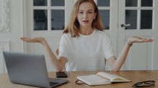 A woman puts her hands palms up as if weighing choices while sitting at her table in front of her laptop.