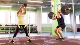 Woman performs kettlebell swing and man performs barbell thruster in gym