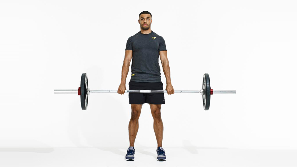 Man facing camera demonstrates how to deadlift, showing midway position, standing upright holding barbell