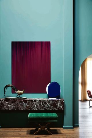 Image of a room with tall walls coated in a cyan-teal paint color. There is a black marble desk on the wall with an emerald green bench seat in front of it. Above the desk hangs a deep magenta painting.