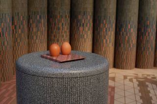 A tile made from dried eggshells is displayed on a grey rounded structure made of tiles as well. On top of the tile, there are two eggshells.