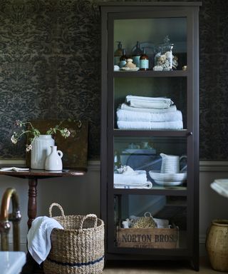 Dark bathroom with tall wooden and glass cabinet, storage and display unit with towels, toiletries and decorative ornaments