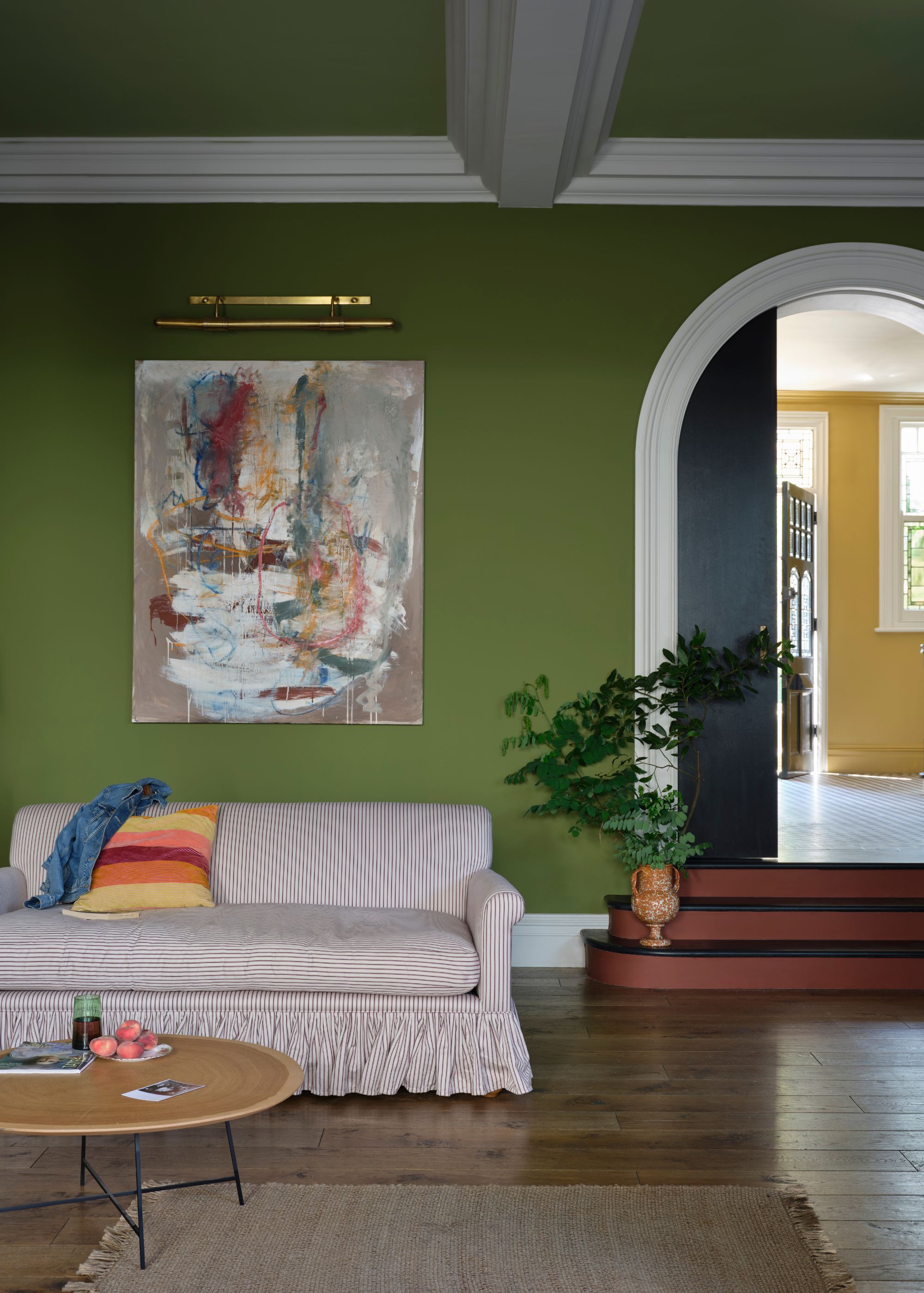 A green living room with white trim details and an arched doorway with steps leading up to it with a vase on the first step filled with foraged foliage; there's also a striped pleated skirt sofa