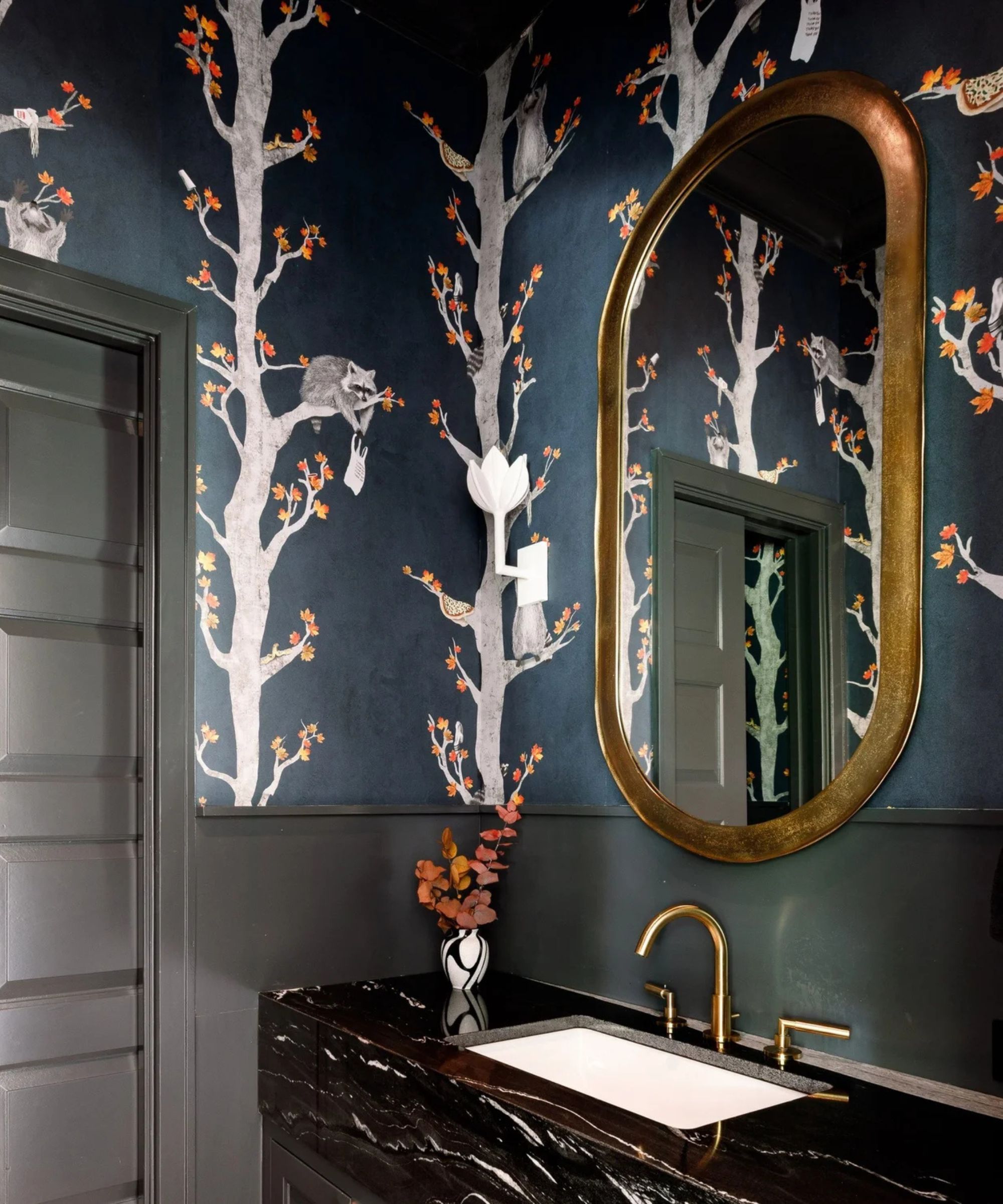 A small bathroom with a black marble sink on a black vanity and moody blue wallpaper depicting trees