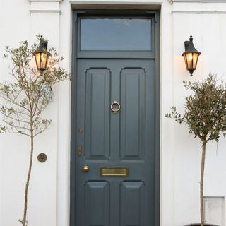 Navy-grey front door flanked by olive trees and lantern garden lights