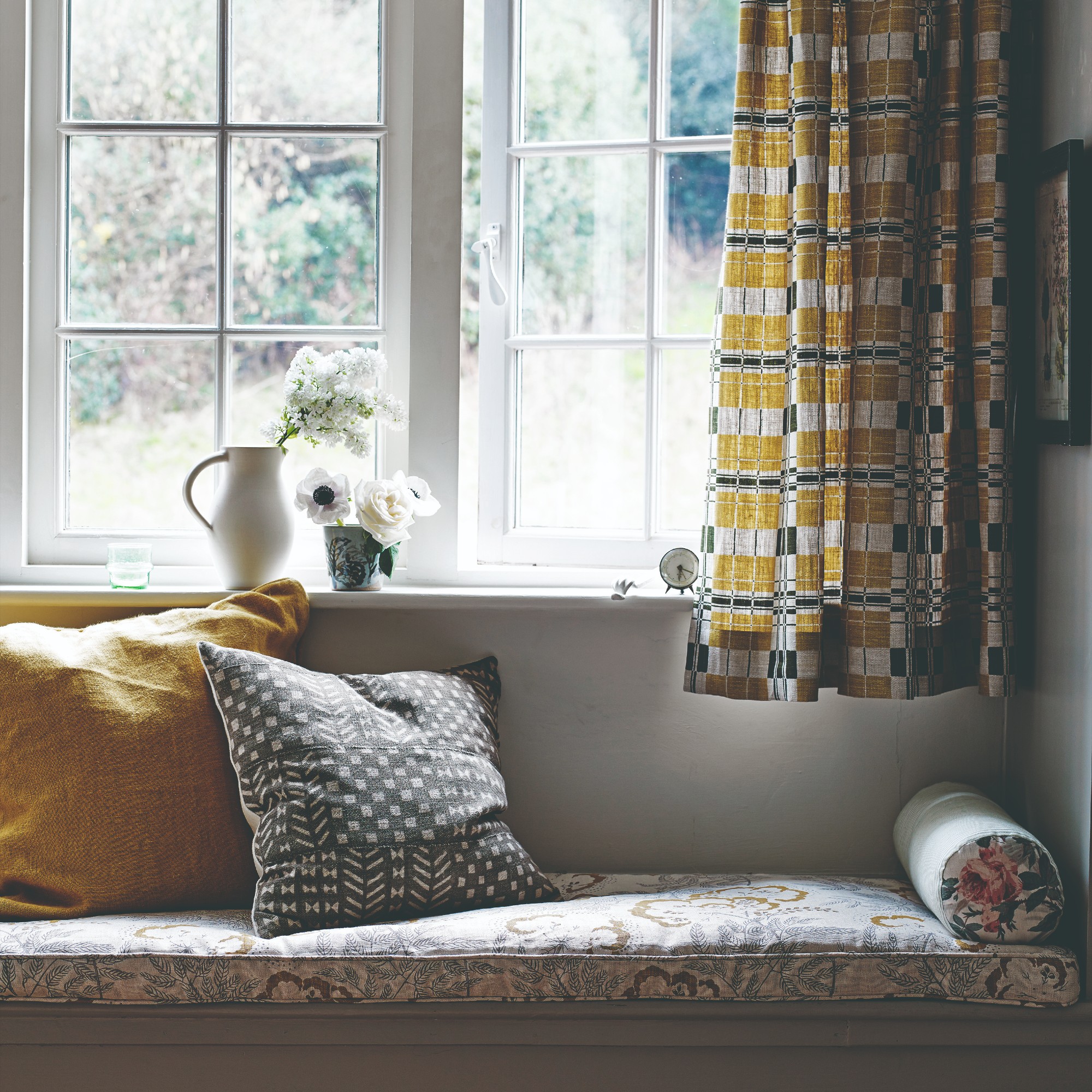 A window seat withb a patterned seat cushion and curtain and the windowsill used for displaying vases of flowers