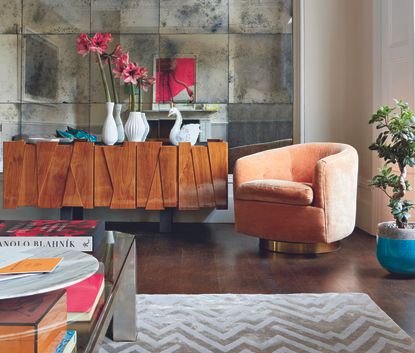  A feature wall of mirrors in a living room reflecting the room, sofa seating and modern furniture and a sideboard clad with a wooden finish and orange and peach accent colours