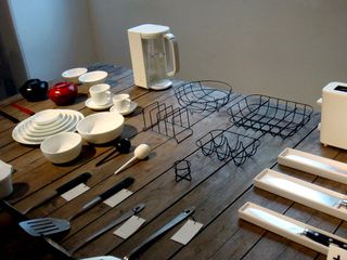 A selection of kitchenware and cookware displayed on a table top, including white plates and bowls, spatulas, knives, a toaster and kettle, two plate holders, a sink drying rack and a fruit bowl.