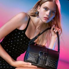 Model holding a Michael Kors black handbag on a pink and blue background