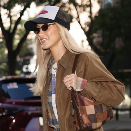 Bella Hadid wearing a brown collared jacket, plaid shirt over a T-shirt, and jeans with a baseball cap, Coach Alter/Ego Slouchy Shoulder Bag, and Ugg Classic Ultra Mini Boots with white socks