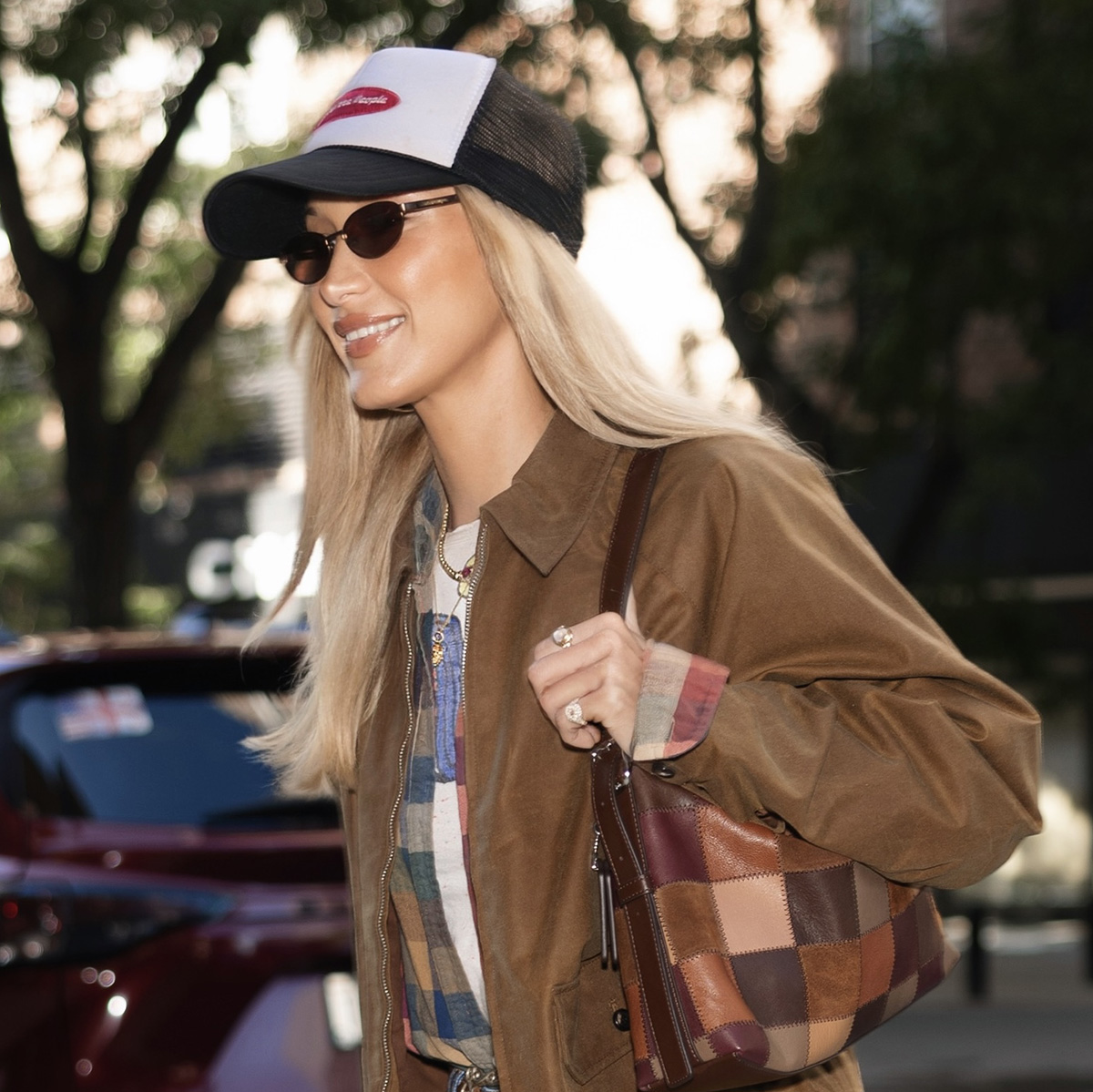 Bella Hadid wearing a brown collared jacket, plaid shirt over a T-shirt, and jeans with a baseball cap, Coach Alter/Ego Slouchy Shoulder Bag, and Ugg Classic Ultra Mini Boots with white socks