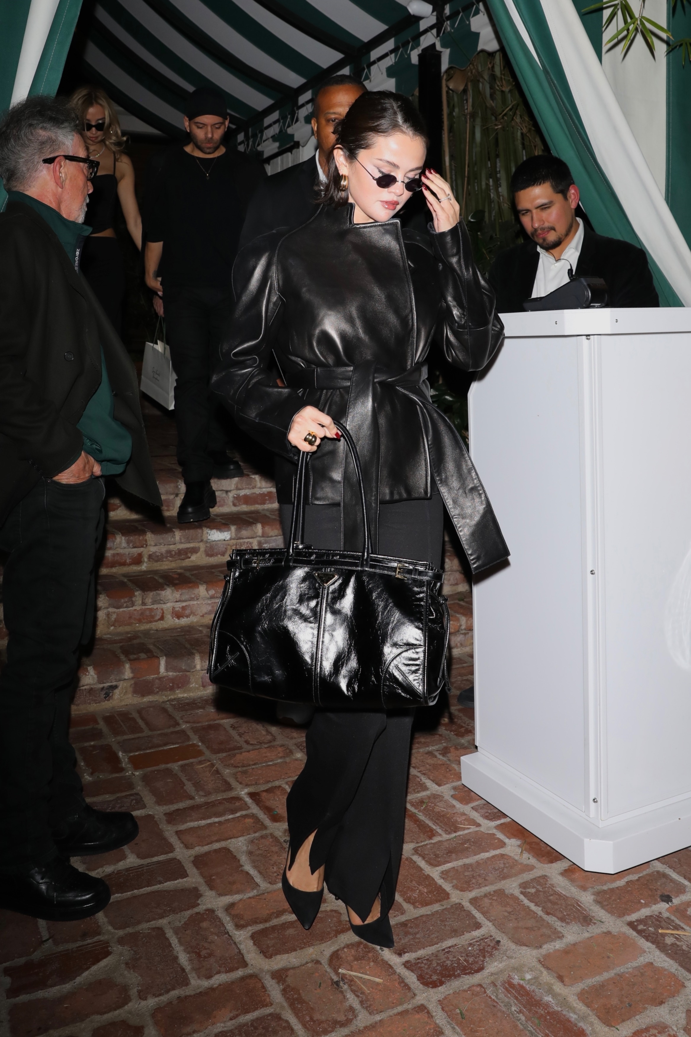 Selena Gomez wears a leather wrap coat, black bag, split-hem pants, and pumps.