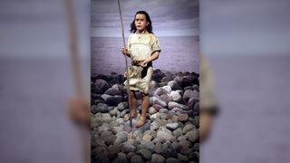 We see a full-body reconstruction of a barefoot teenager with dark, shoulder-length hair standing on the rocky shore. He holds a fishing pool and wears a leather tunic.