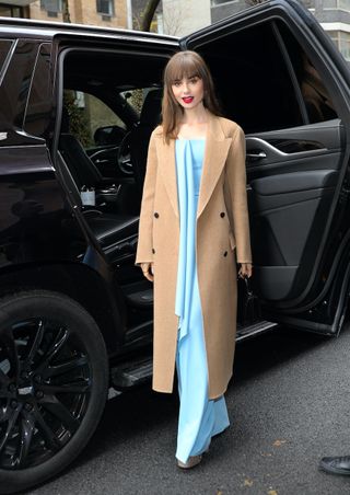 Lily Collins leaving an event in New York City on December 12, 2022 wearing a long camel coat, a light blue draped top and pants from Del Core, a Cartier mini bag, and Valentino crystal-embellished platform heels