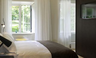 Inside a bedroom with a large bed made up with white linen, a brown throw, white draping curtains and a window.