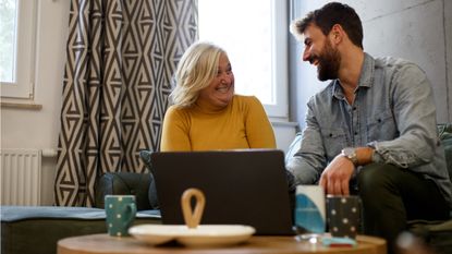 A mother and adult son smile and laugh while sitting in front of a laptop on a coffee table.