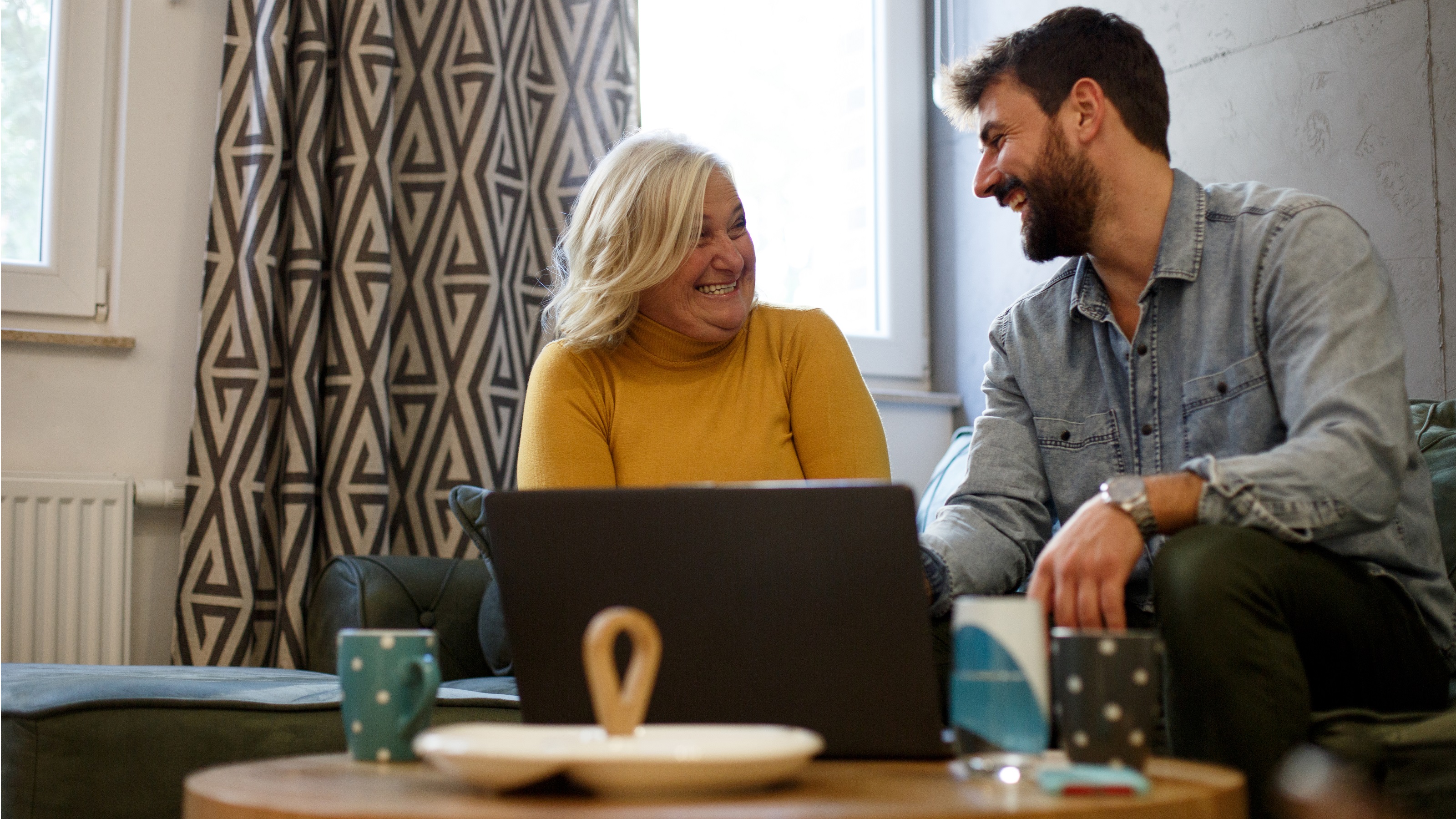 A mother and adult son smile and laugh while sitting in front of a laptop on a coffee table.