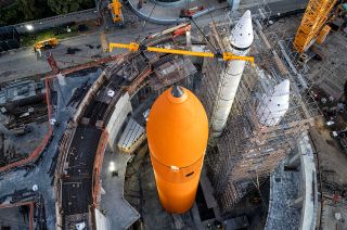 a large cylindrical orange fuel tank stands next to two large white rocket boosters, which are surrounded by scaffolding.