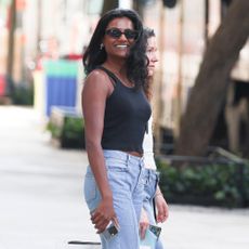 Simone Ashley stands outside wearing a black tank top with blue flared jean and black sunglasses.