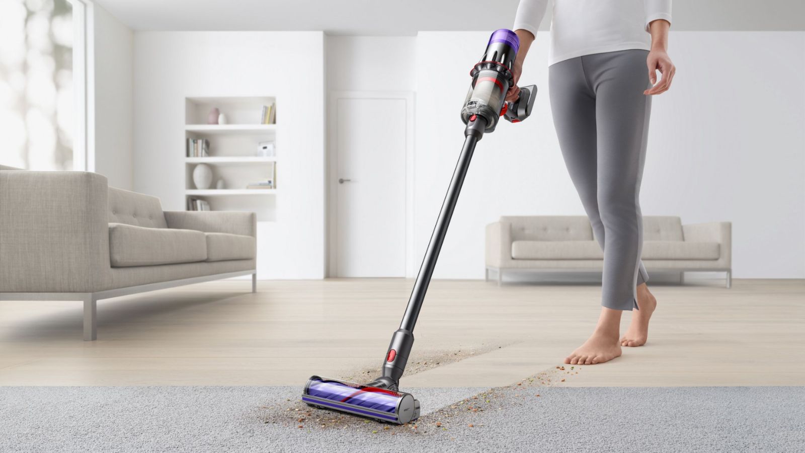 A woman using the Dyson V9 Motorbar Cordless vac. Her head is not visible, and she is wearing a white long-sleeve top, grey trousers, and her feet are bare. She is using the vacuum between hardwood flooring and grey carpet, in a large living room with cream couches, white walls, and in-built white shelving.