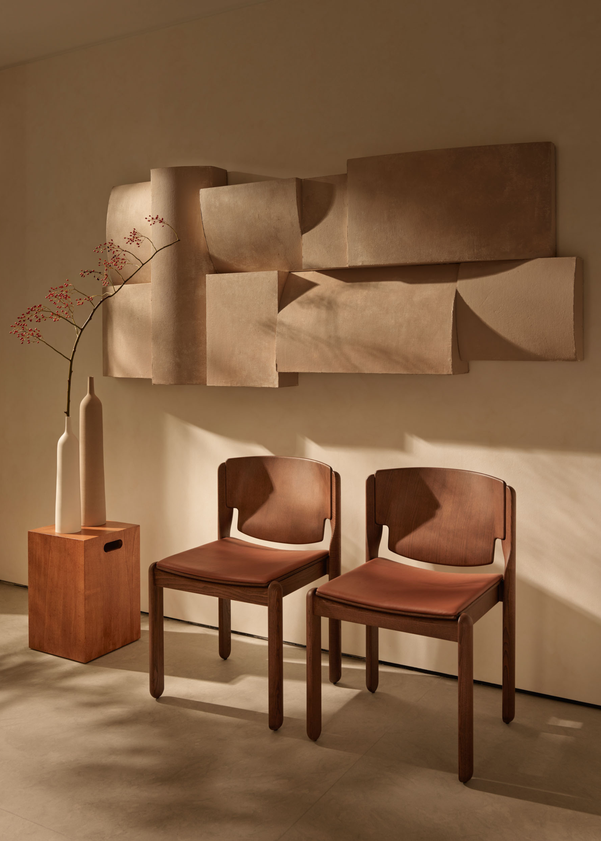 An off-white living room corner with leather chairs, a wood side table with a pair of beige vases and a structural wall hanging