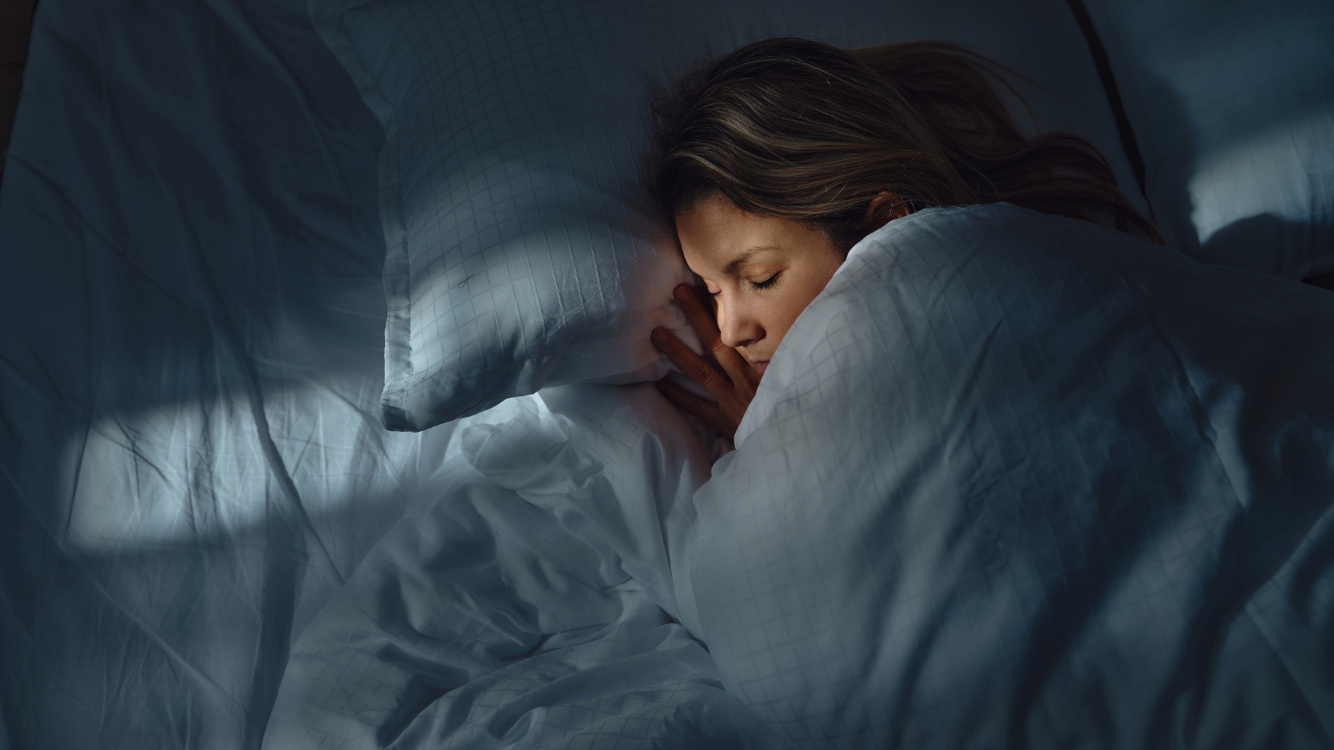 A woman sleeping on her side at night in a bed with all white bedding.