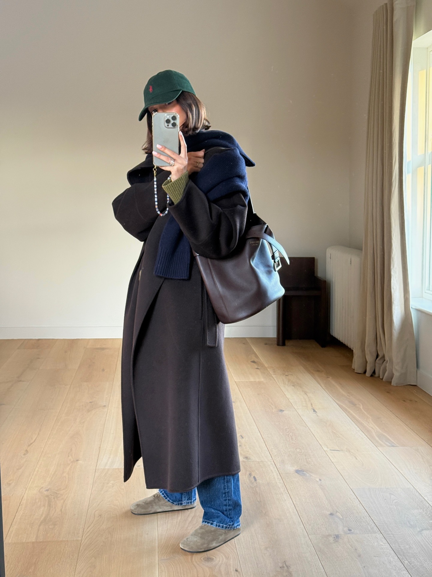 Woman wearing jeans, flat suede mules, a long coat, and a baseball cap.