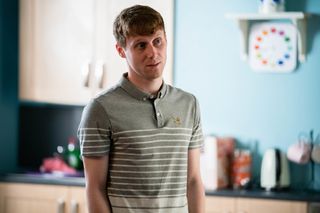 Jay Brown wearing a grey t-shirt and standing in the kitchen in EastEnders 