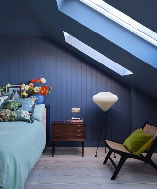 Blue painted bedroom with sloped ceiling, skylights, blue bedding and floral cushions, lounge chair, lantern floor lamp, bedside table