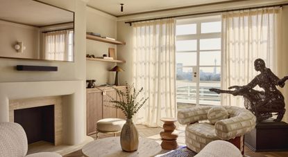 A sun-filled living room decorated with beige and pale wood-tinted furniture and a metal statue of a woman put on a dark wood podium.