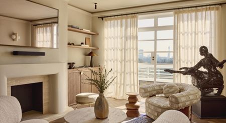 A sun-filled living room decorated with beige and pale wood-tinted furniture and a metal statue of a woman put on a dark wood podium.