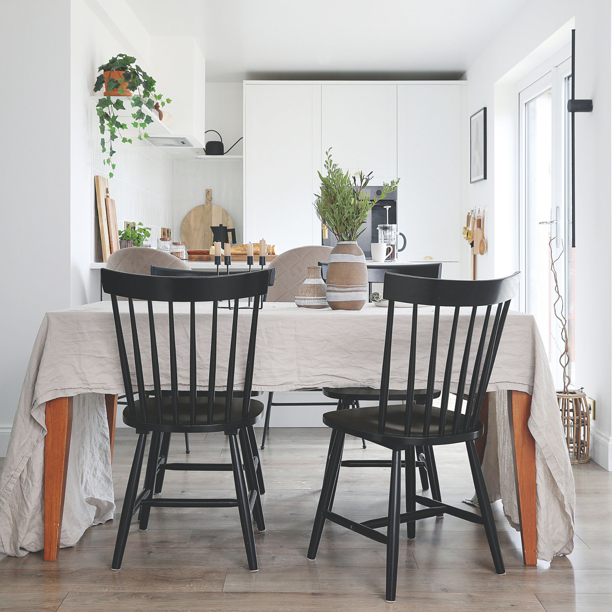 A dining area with black chairs and a rectangular table covered with a linen tablecloth connected to a white kitchen