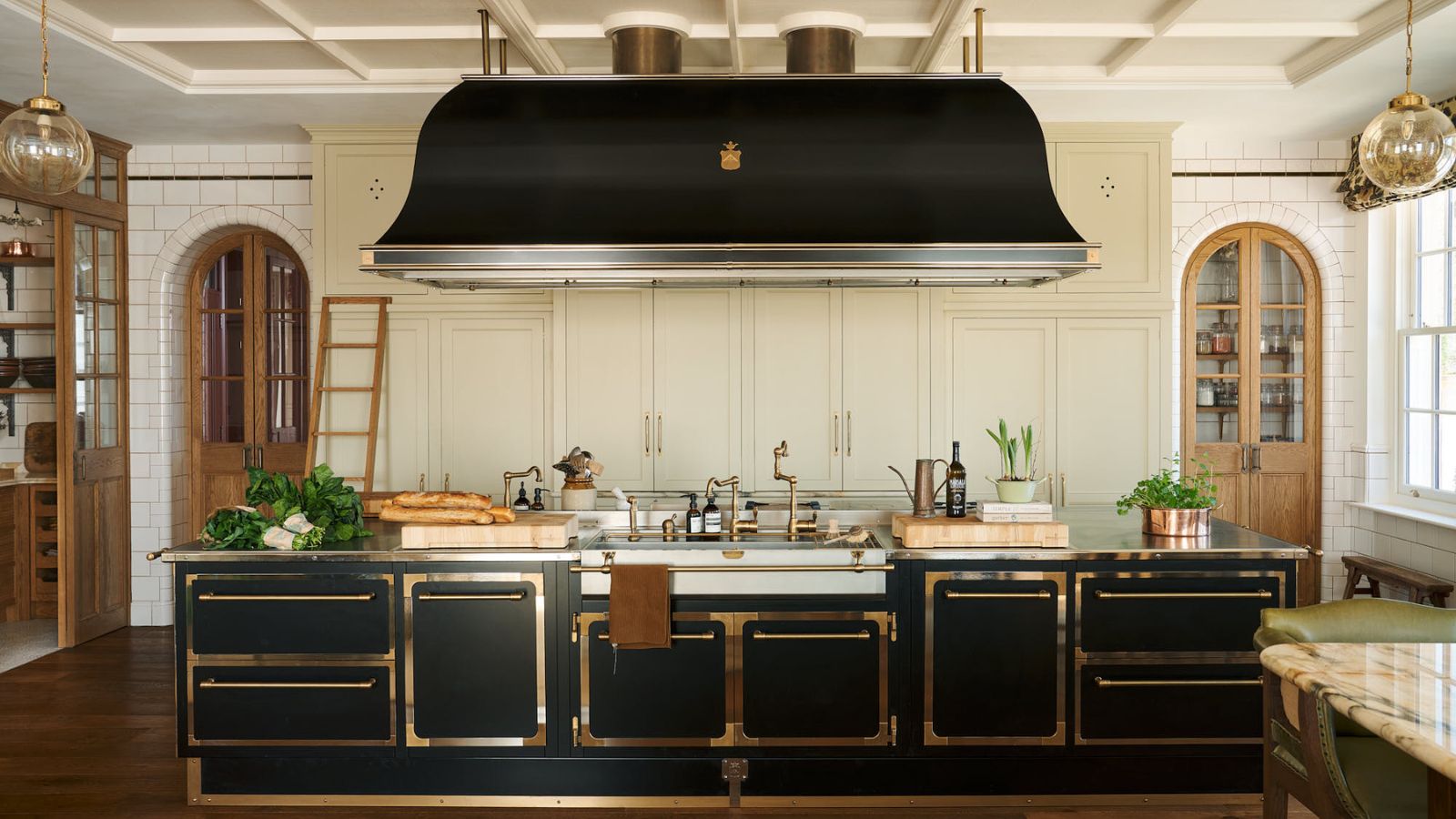 Large black and cream kitchen with vintage touches. Its cabinets have brass borders, and the large black extractor hood above the island is an eye-catching and unusual focal point that makes this kitchen feel very grand and 1920s salloon style