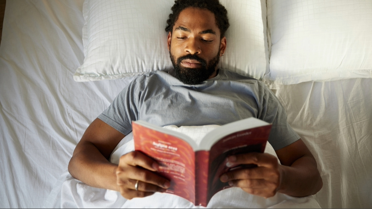 A man laying in bed with white sheets reading a book with red cover.