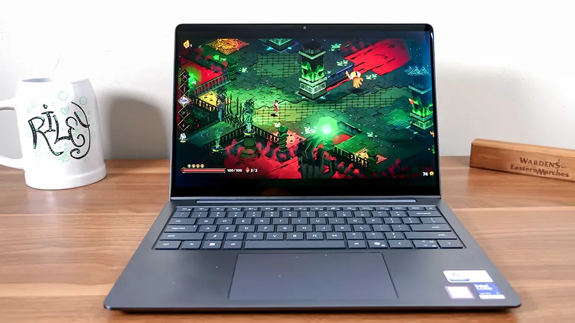 A laptop on a wooden desk displays a vivid top-down action game with neon green and red combat scenes; a white mug and wooden nameplate sit nearby.