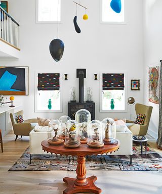 white living room with woodburner, sculptural armchairs, white sofa, black mobile, round wooden table with glass domed birds and colorful rug