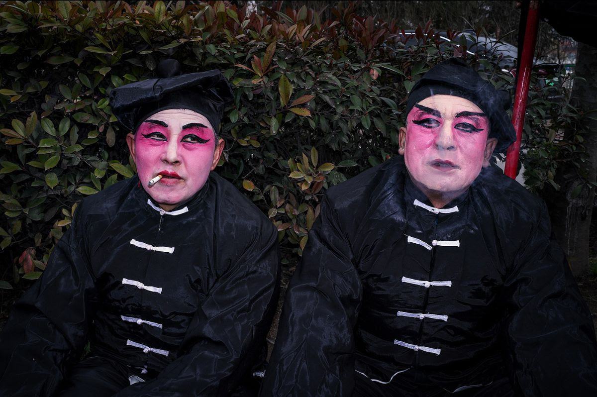 Two performers with vivid pink face makeup and dramatic black eye markings wear matching black traditional costumes with white toggle closures against a backdrop of dark foliage, one smoking a cigarette.&amp;#57430;&amp;#57403;&amp;#57595;&amp;#57593;&amp;#57550;