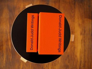 Two books with a red cover say 'Donald Judd Writings'. They're photographed on a black stool.