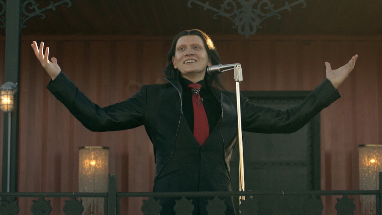 Calypso in black suit and red tie holding his arms out while announcing tournament details in Twisted Metal Season 2