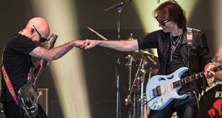 Joe Satriani and Steve Vai fistbump onstage during their show at the Valley Center, California in May 2024. They have their signature Ibanez guitars. Satch has the Chrome Boy JS1. Vai is playing his Daphne Blue PIA in Powder Blue
