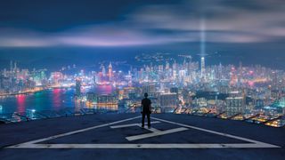 A person stands on a helipad at night, overlooking a vibrant, illuminated city skyline across the water, with a dramatic sky above