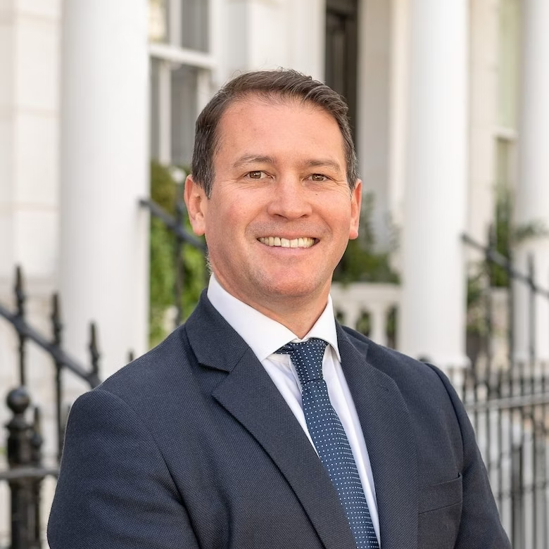 male with short dark hair wearing navy suit, white tie and blue tie stood outside white houses with column and railings
