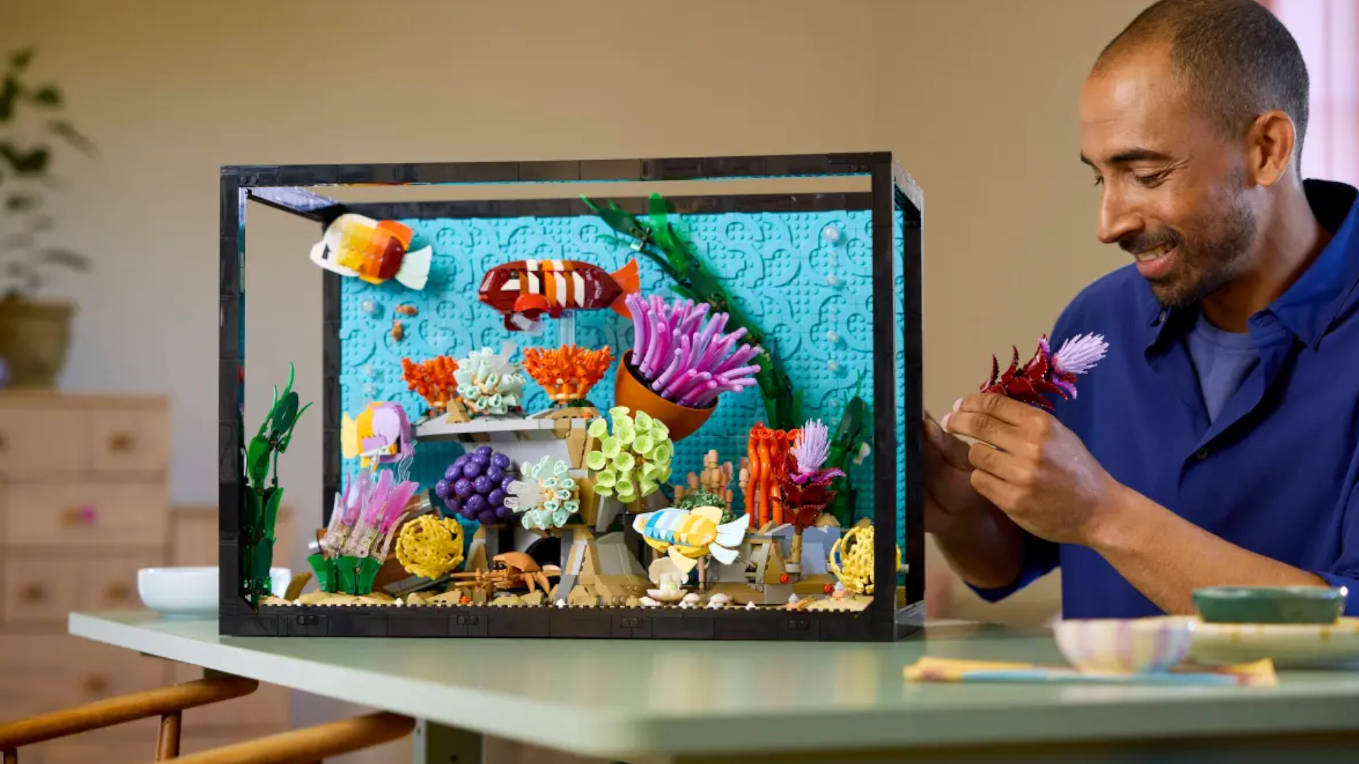 A man smiles while placing a piece onto the Lego Tropical Aquarium kit, which is mostly assembled on a table