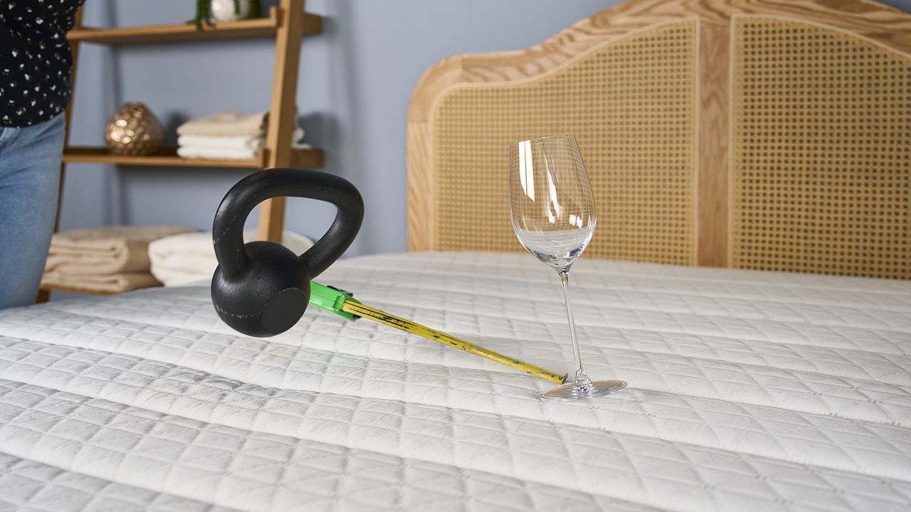 A person drops a 10lb weight on to the surface of the Helix Dawn Mattress, 10 inches away from a wine glass to test motion isolation. The wine glass is wobbling from the impact
