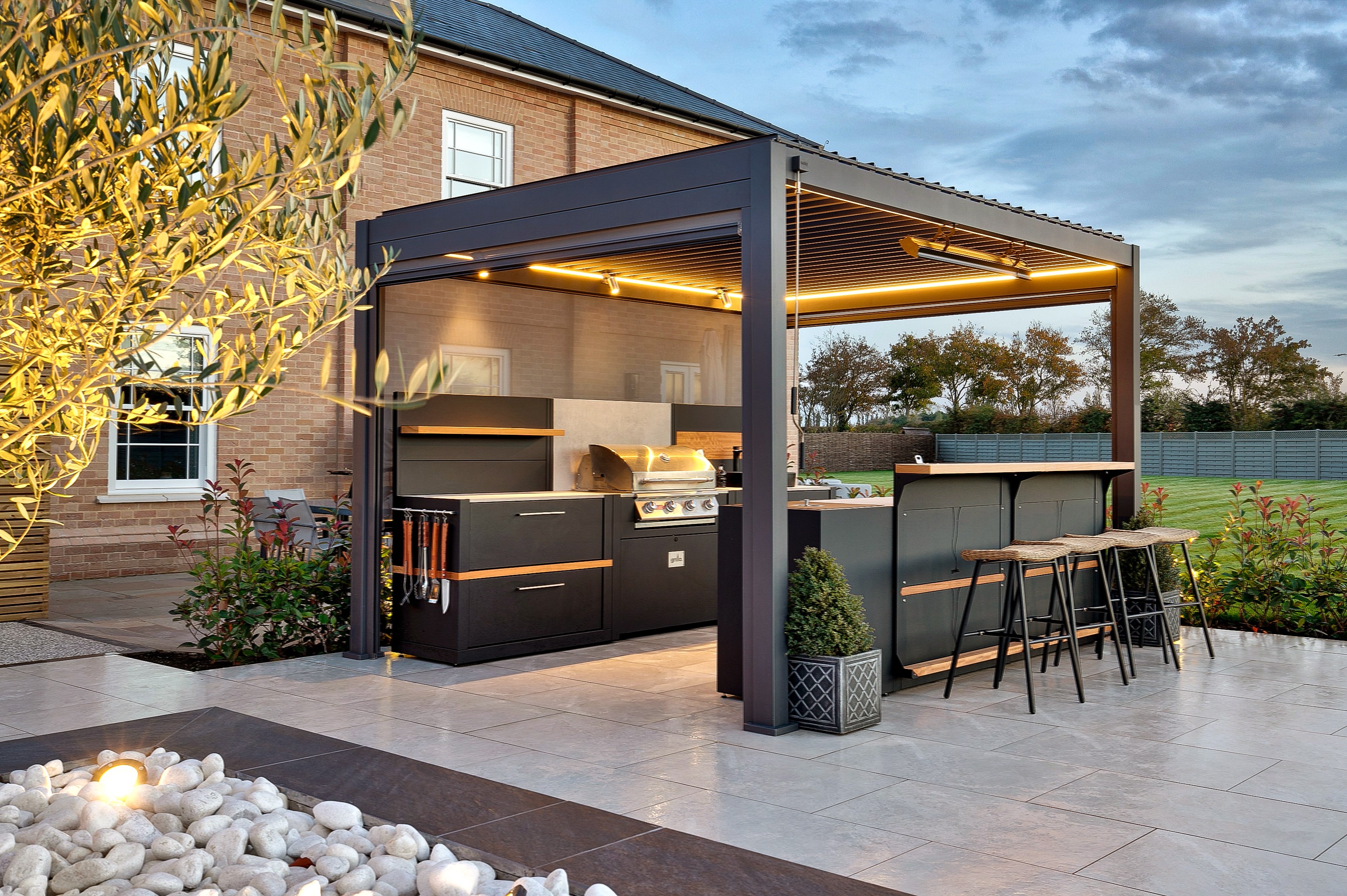 An outdoor space with a kitchen within a pergola, lit up with recessed lights