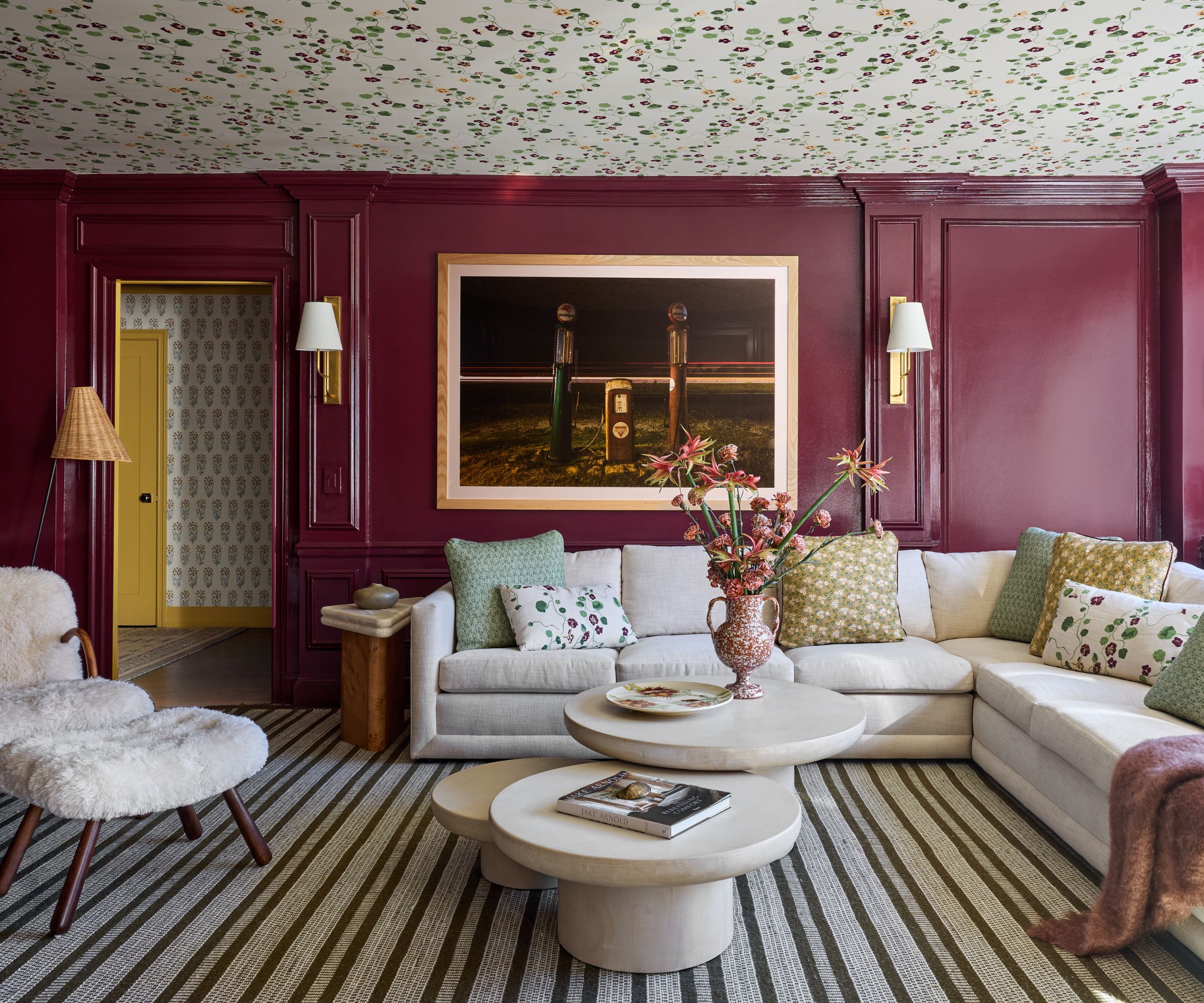 A living room with burgundy paneled walls in a gloss finish and a floral wallpapered ceiling. A striped carpet, a large cream sectional with green, yellow, and floral cushions, a modern three-piece coffee table, artwork on the wall with two sconces, and a shearling accent chair.