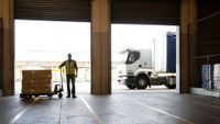 A worker wearing a yellow vest stands looking into an empty warehouse with a cart full of food boxes next to him and a truck in the background