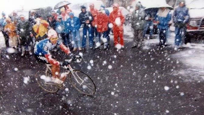 A cyclist climbs a mountain in heavy snow with a woolly hat on